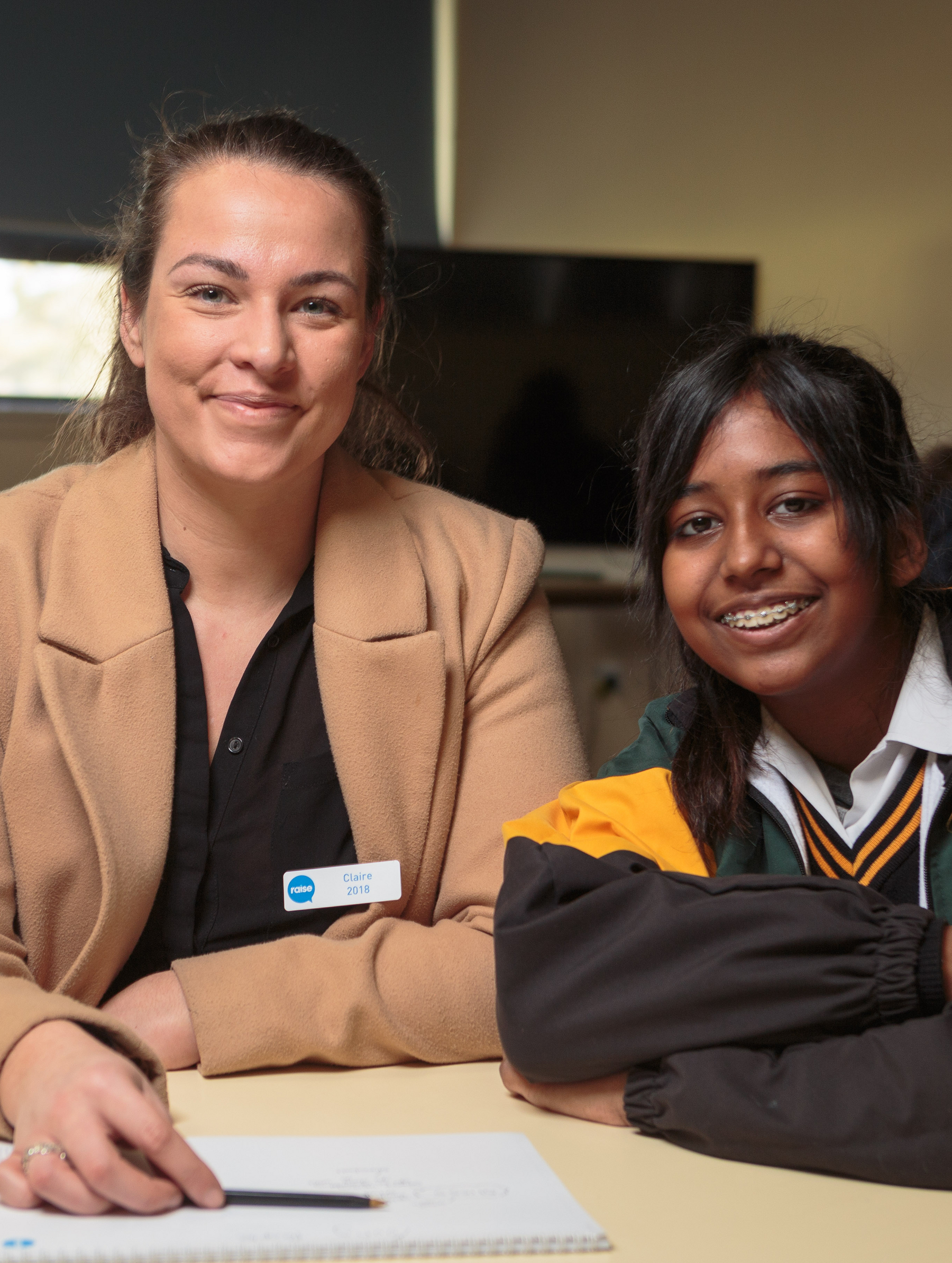 Raise Foundation mentoring program. Two people seated at a table, one wearing a tan coat and name badge, the other in a school uniform, with a notebook and pen on the table in front of them.