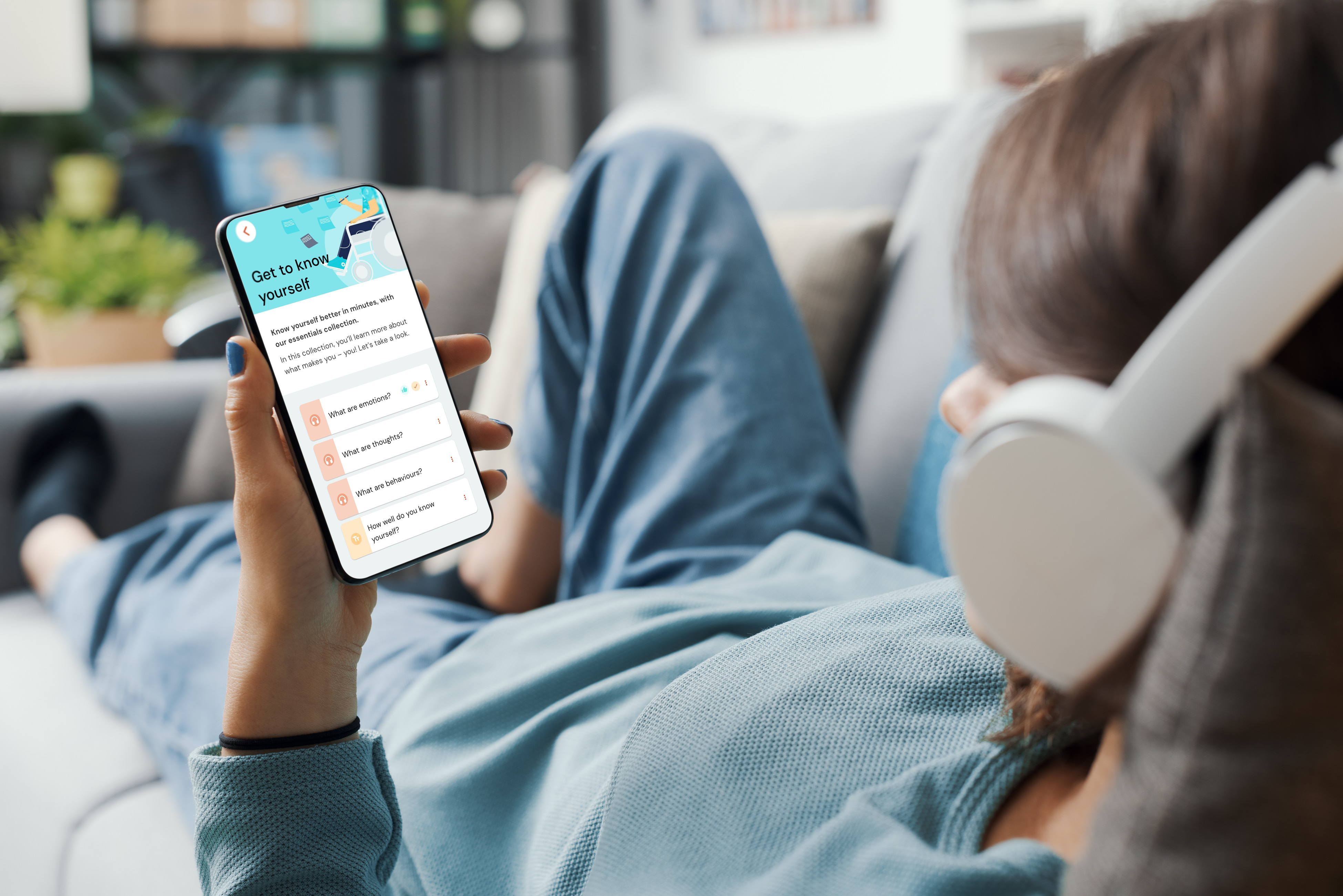 Person relaxing on a sofa wearing headphones, holding a smartphone displaying a self‑reflection app with colourful question cards.