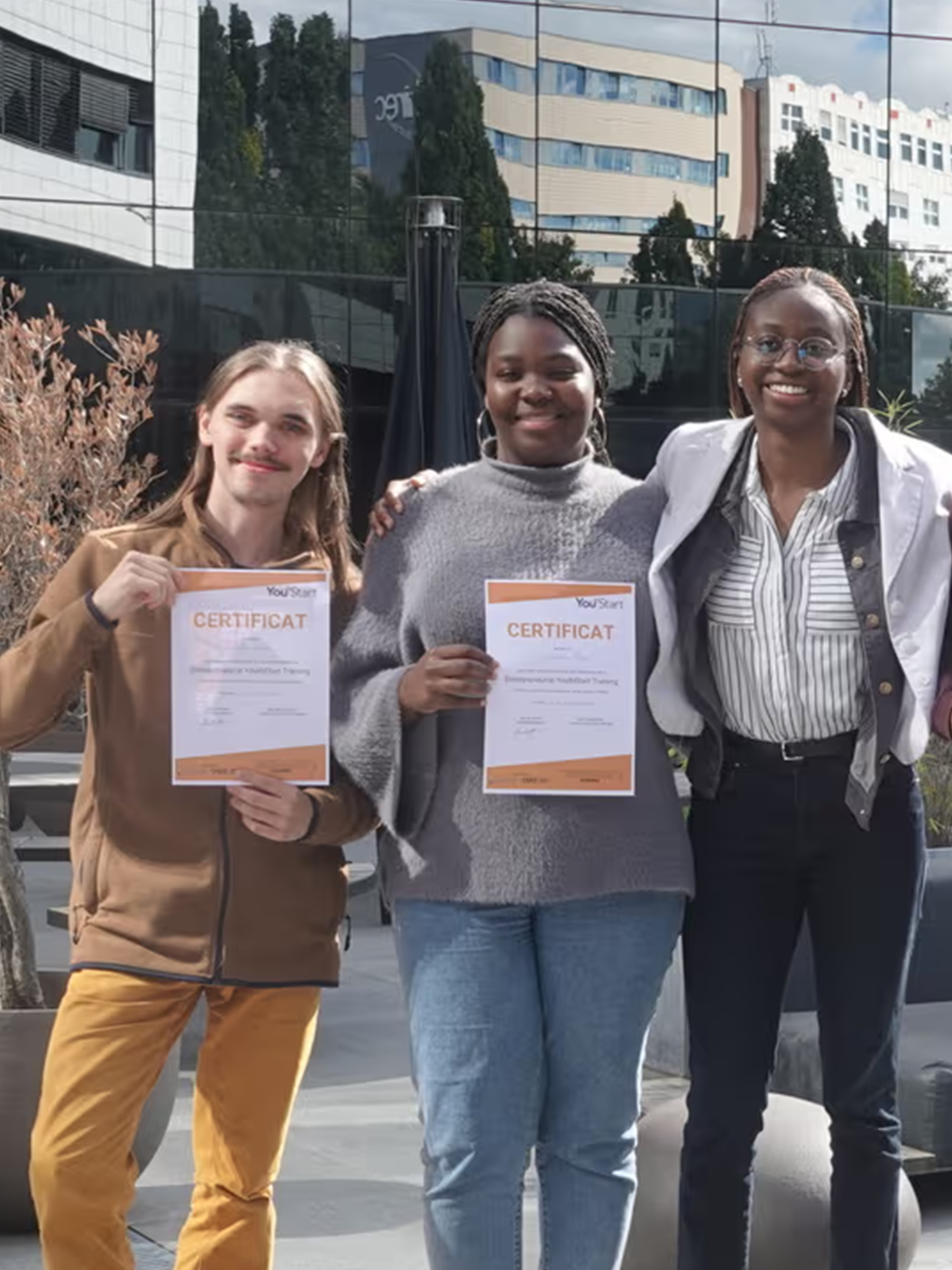 Youth Start Belgium Three people standing outdoors holding printed certificates at an urban courtyard with modern buildings behind them.